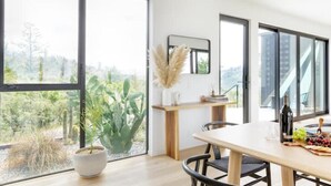 A modern dining area with large windows, a wooden table, and a view of greenery outside.