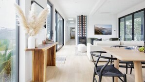 A modern interior with a wooden dining table, white sofas, and large windows offering a view of the outdoors.