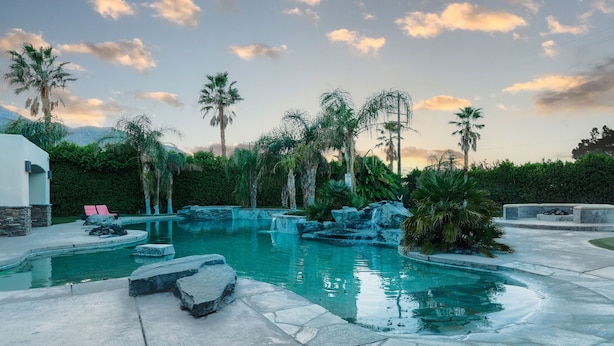 A swimming pool with a waterfall feature, surrounded by palm trees and a stone deck. - The Galvan Estate (Palm Springs)