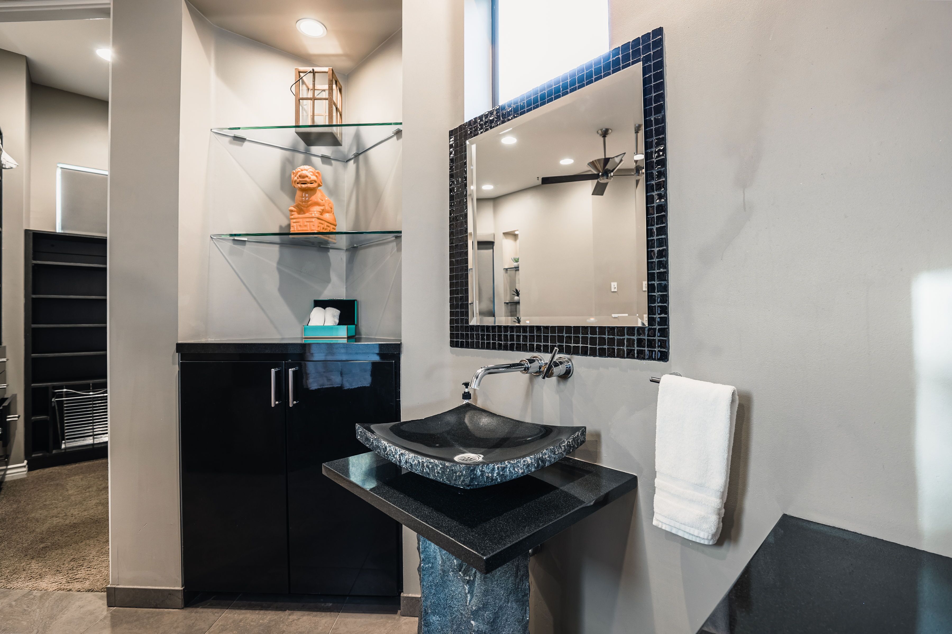 A modern bathroom with a black granite sink, a glass shelf with a decorative item, and a mirror with a black tile backsplash.