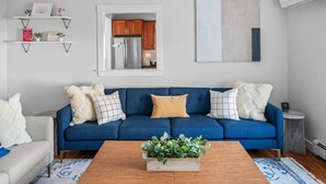 Ein Wohnzimmer mit einem blauen Sofa, einem Holztisch mit einer Pflanze und einer durchgehenden Tür sichtbaren Küche.