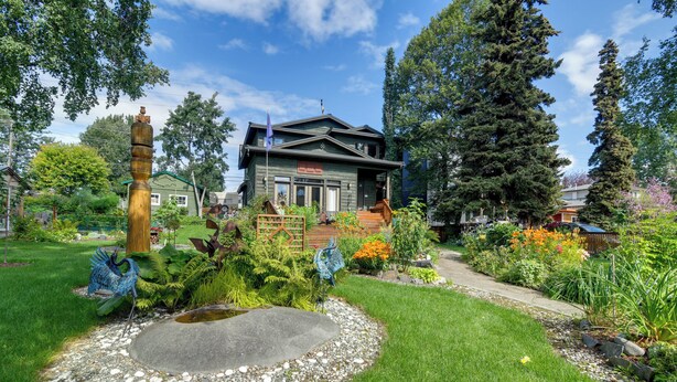 A two-story house with a front deck, surrounded by a well-maintained garden with various plants and a decorative metal sculpture.