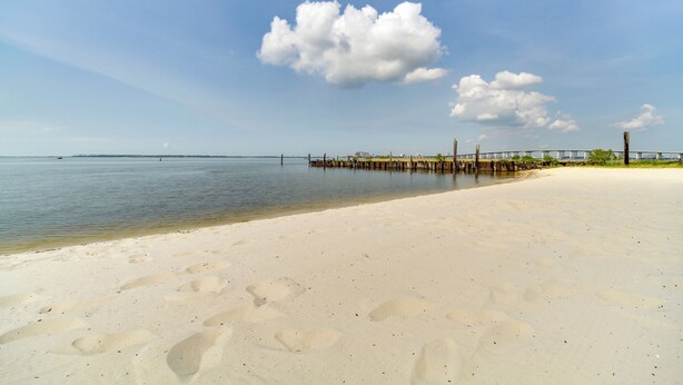 Een strand met een houten steiger die zich uitstrekt over rustig water, een heldere lucht en voetafdrukken in het zand.