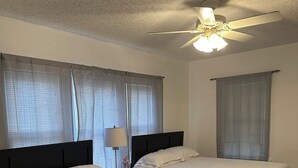 Una stanza con due letti, un ventilatore a soffitto e ampie finestre con tende leggere.