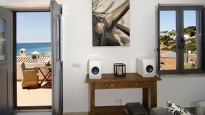 A room with a view of the sea through an open door, a painting of a ship on the wall, and a wooden table with two speakers.