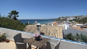 A terrace with wicker furniture, a table with a vase of flowers, and a view of the sea and a hillside town.