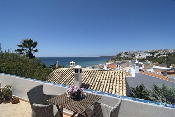 A terrace with wicker furniture, a table with a vase of flowers, and a view of the sea and a hillside town.