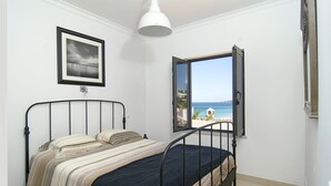 A bedroom with a bed, a framed picture on the wall, a window with a view of the beach, and a ceiling light.