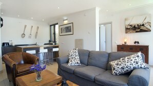 A modern living room with a grey sofa, a wooden coffee table, and a brown leather armchair. There is a kitchen area with a bar counter and a dining table with bar stools.