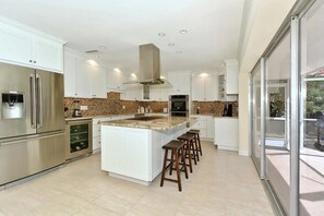 Une cuisine moderne avec des meubles blancs, un îlot central doté de tabourets de bar, des appareils en acier inoxydable et une grande fenêtre offrant une vue sur l’extérieur.