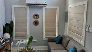 A living room with a grey sofa, a coffee table with a plant, and a blue rug. There are two windows with blinds, an air conditioning unit, and a wall-mounted clock.