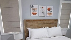 A bedroom with a wooden headboard, white bedding, and a teal throw blanket. There are three wall-mounted artworks above the bed.