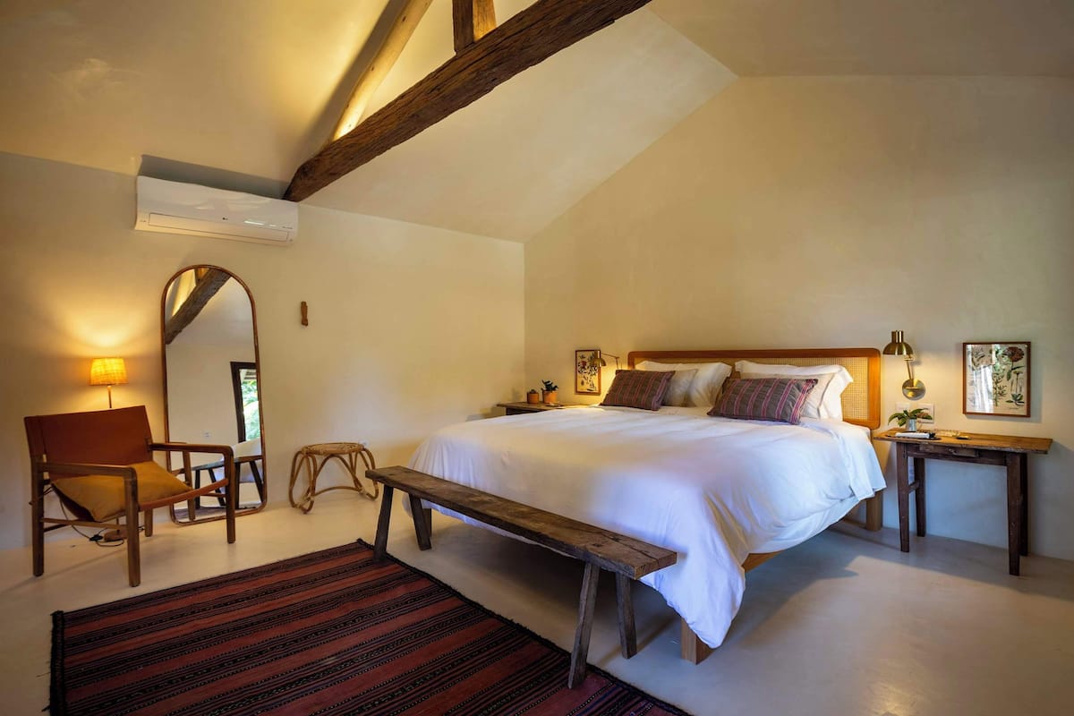 A bedroom with a large bed, a wooden bench, a chair, a bedside table, and a mirror.