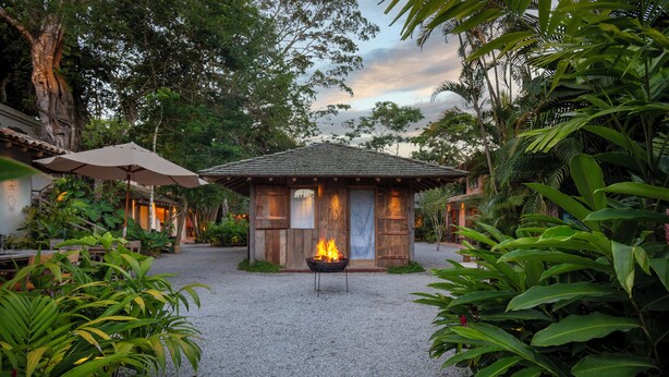 A rustic wooden structure with a fire pit in the center, surrounded by lush greenery and a gravel path. - Na Praça Hotel (Porto Seguro)