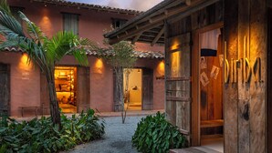 A rustic building with a wooden door, a small garden with tropical plants, and a visible interior with shelves and furniture. - Na Praça Hotel (Porto Seguro)