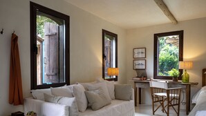 A cozy room with a white sofa, a wooden desk, and a chair. There are windows with wooden shutters, a lamp, and a small table with a plant. - Na Praça Hotel (Porto Seguro)