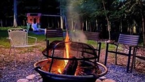 Ein Lagerfeuer mit einem Metallrost, umgeben von Steinen, in einer bewaldeten Gegend mit Lichterketten darüber.