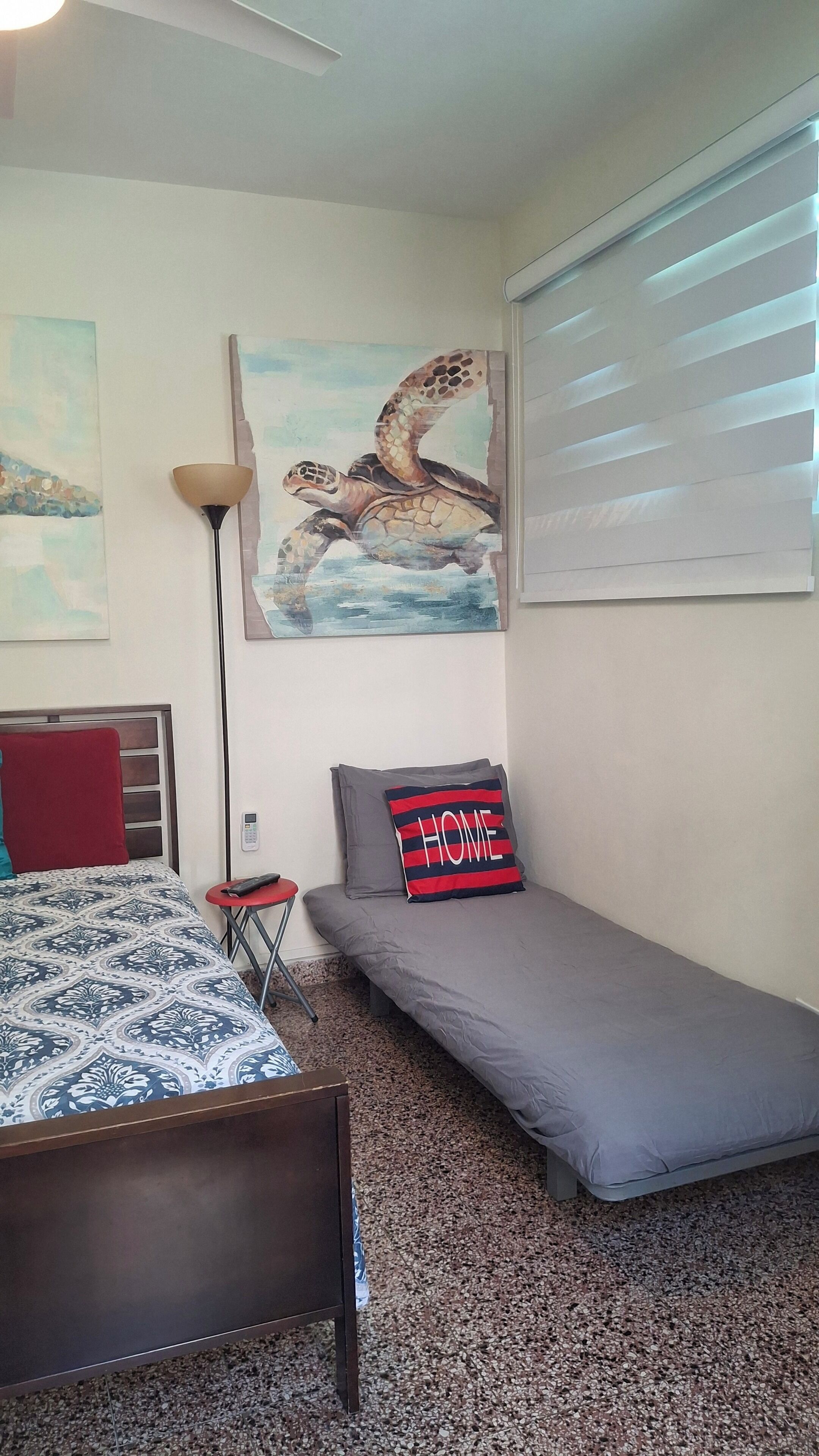 Un dormitorio con una cama, una mesita de noche pequeña, una lámpara, un cuadro colgado en la pared y una ventana con persianas, además de un colchón gris.