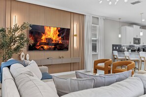 Ein modernes Wohnzimmer mit Fernseher, der ein Feuer zeigt, einer Couch, einem Couchtisch und einem Essbereich mit Stühlen.