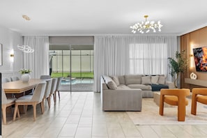 A modern living room with a grey sofa, a dining table with chairs, a chandelier, and a large window with curtains. - New Home 8 Bedrooms and 5 Bathrooms At Storey Lake (Kissimmee)