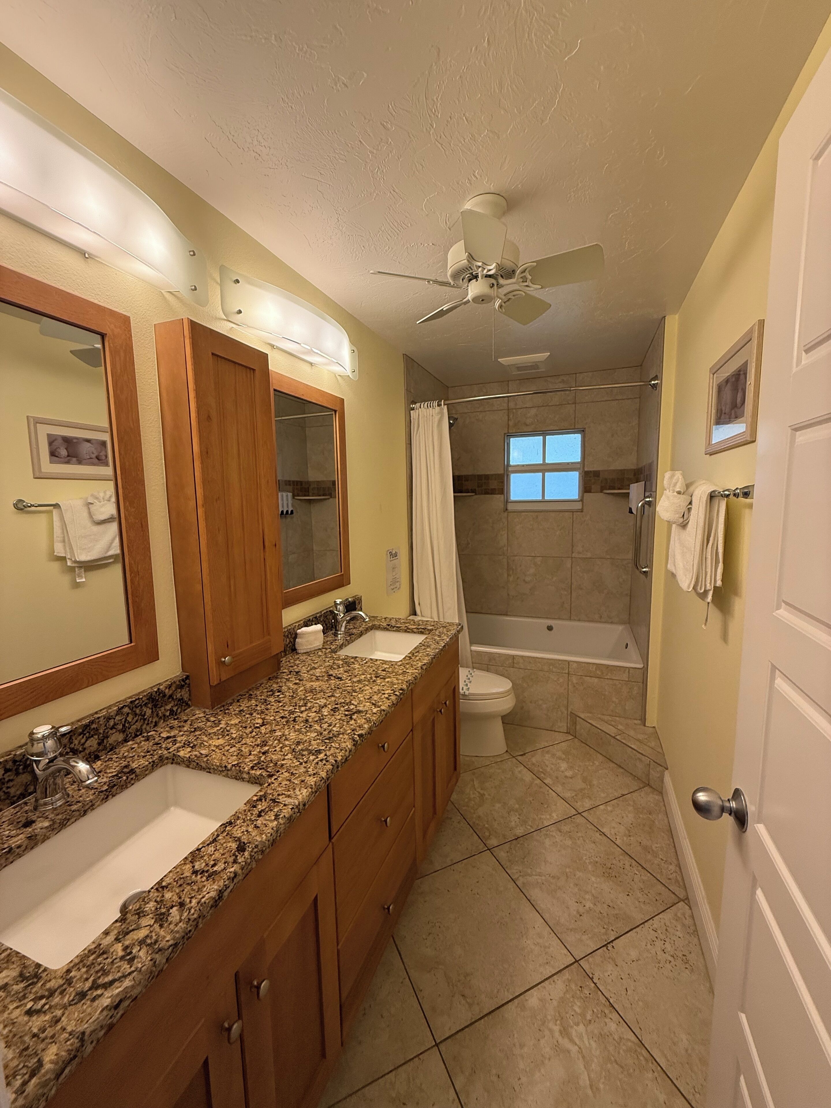 A bathroom with a granite countertop, double sinks, a shower, and a ceiling fan.