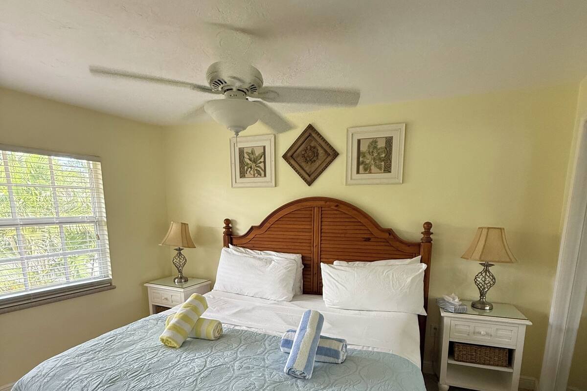 A neatly made bed with a blue quilt, two white pillows, and two folded towels. There is a wooden headboard, a nightstand with a lamp, and a wall with two framed pictures.