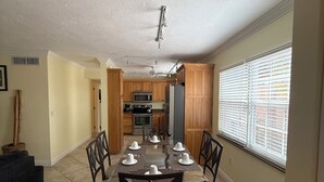 A dining area with a table set for six, wooden cabinets, a kitchen in the background, and a window with blinds. - Siesta Key Inn (Siesta Key)