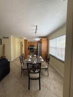 Comedor con una mesa lista para seis comensales, armarios de madera, una cocina al fondo y un ventanal con persianas.