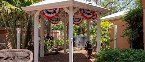 A gazebo with white pillars and red, white, and blue drapes, surrounded by lush greenery and a brick patio.