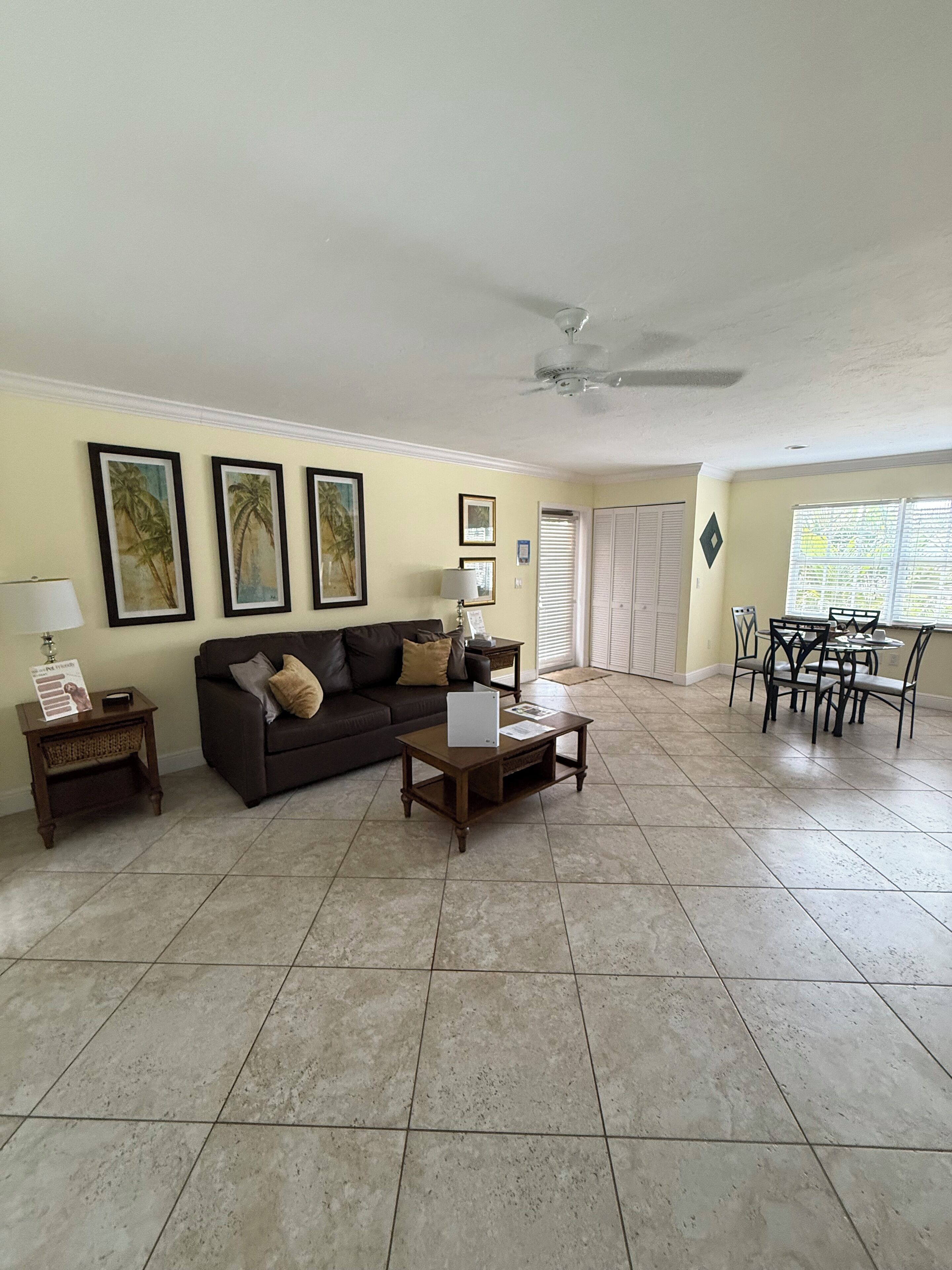 A living room with a sofa, coffee table, and dining area with chairs. There are framed pictures on the wall and a ceiling fan.