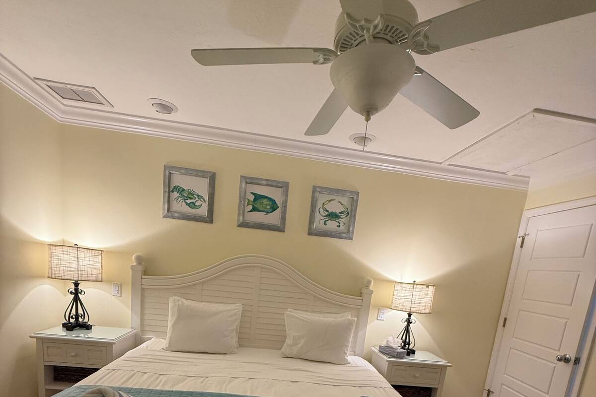 A neatly made bed with a teal bedspread, two rolled towels, and a ceiling fan above. There are framed pictures on the wall and a door to the right.