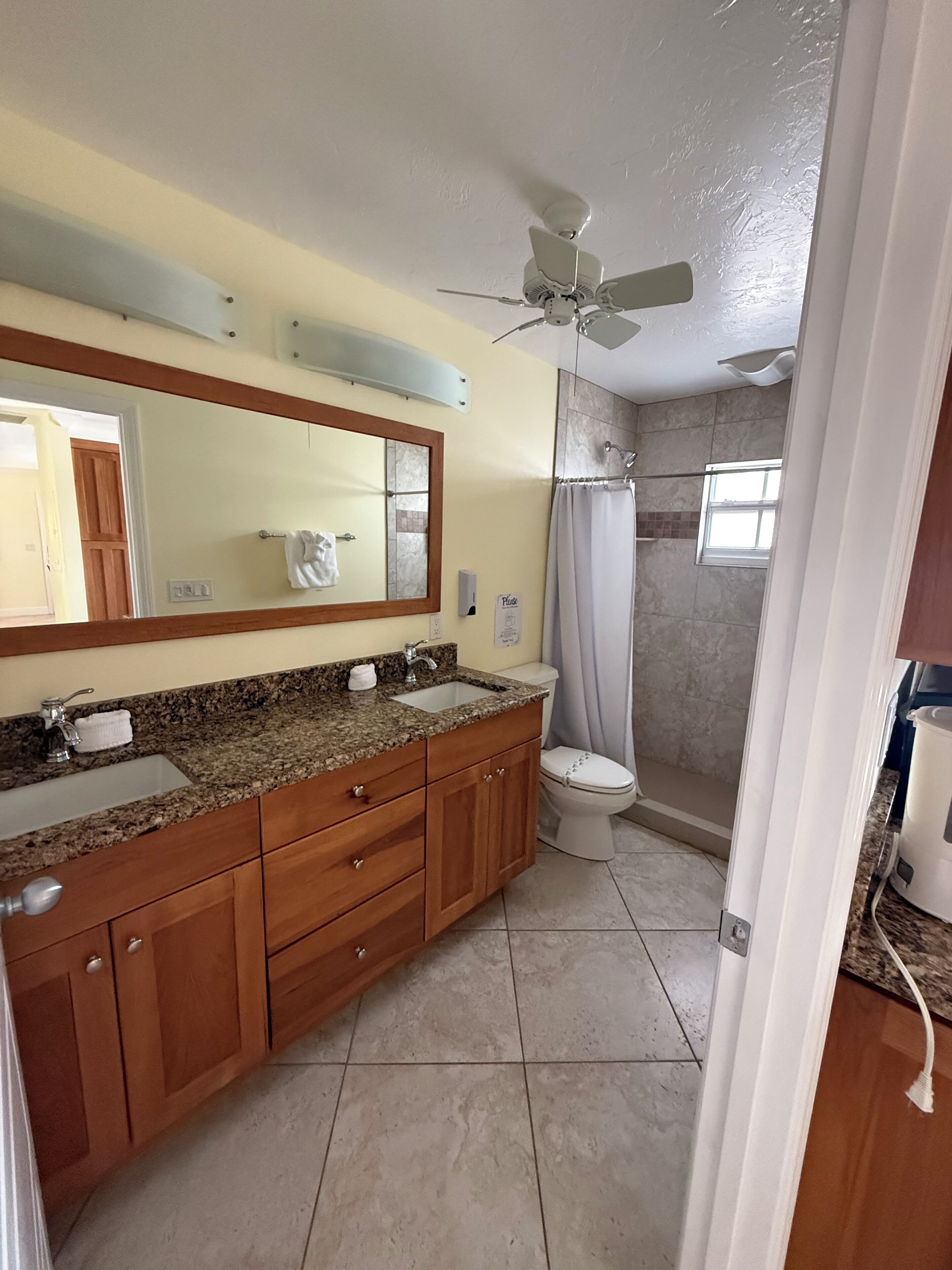 Un baño con encimera de granito, armarios de madera, inodoro blanco, ducha con cortina y ventilador de techo.