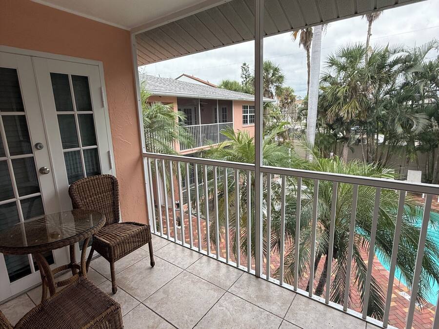 Un balcon agrémenté de meubles en osier, d’une table en verre et offrant une vue sur une piscine et des palmiers.