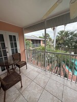 Un balcón con mobiliario de mimbre, una mesa de vidrio y vista a una piscina y palmeras.