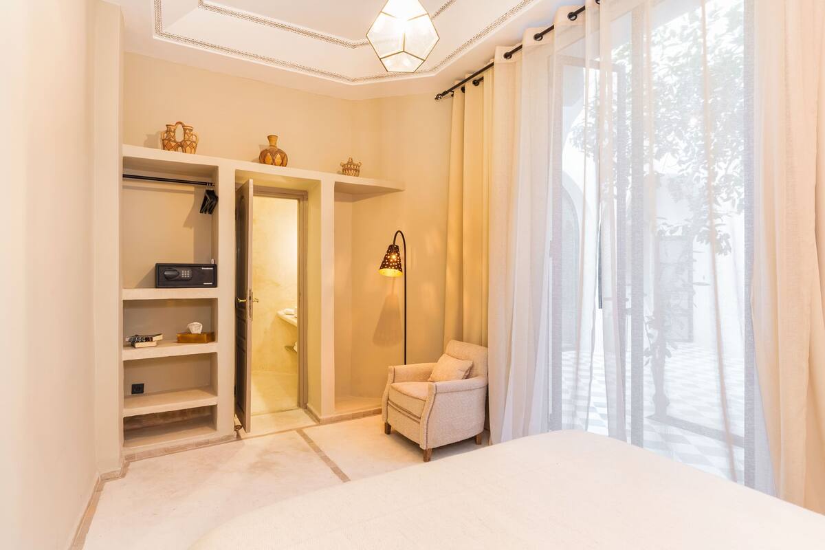 A bedroom with a bed, a chair, a wardrobe, and a window with curtains.