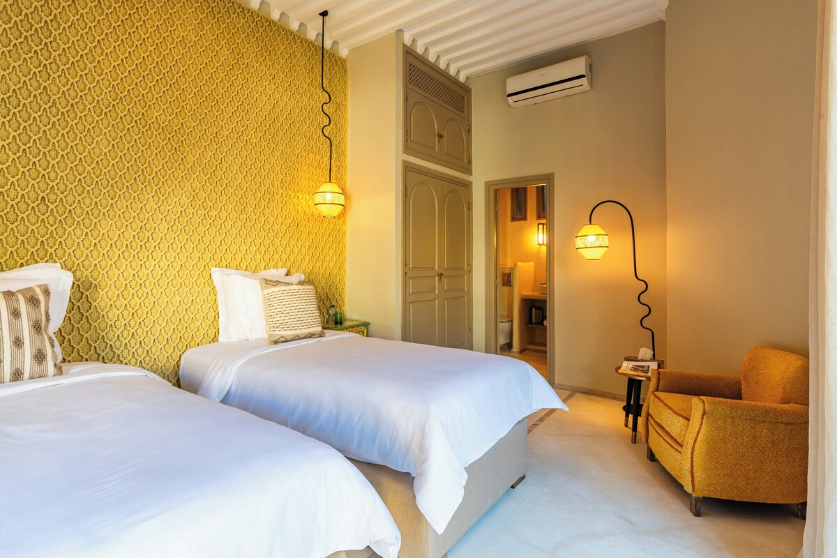 A hotel room with two beds, a small table, a chair, and a door leading to a bathroom.