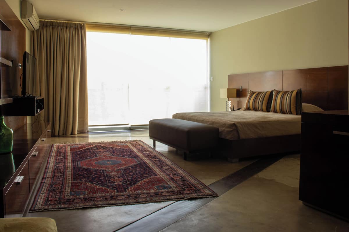 A hotel room with a bed, a desk, a TV, a window with curtains, and a patterned rug.