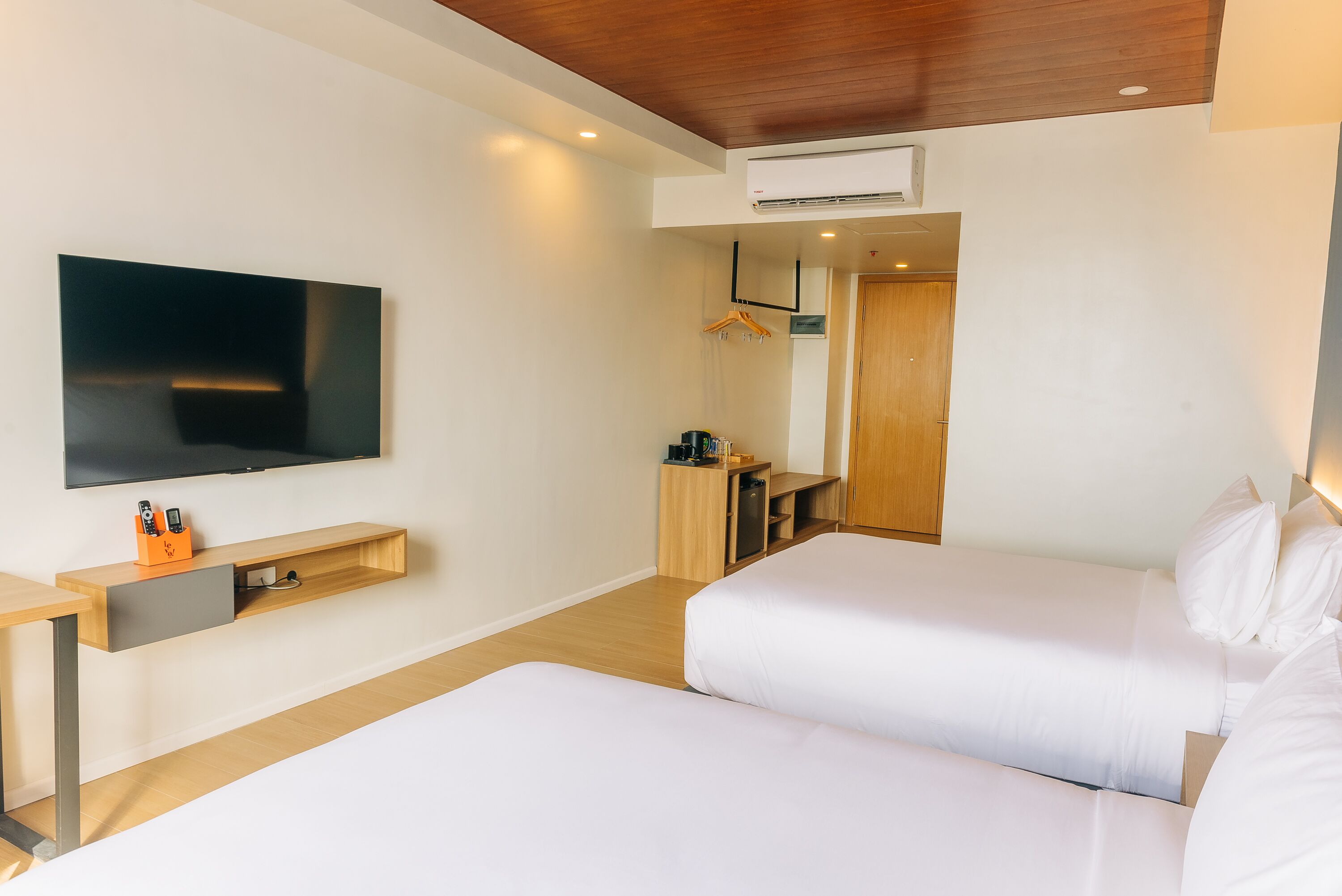 A modern hotel room with a flat-screen TV mounted on the wall, a wooden desk with a drawer, a bed with white bedding, and a wooden ceiling.