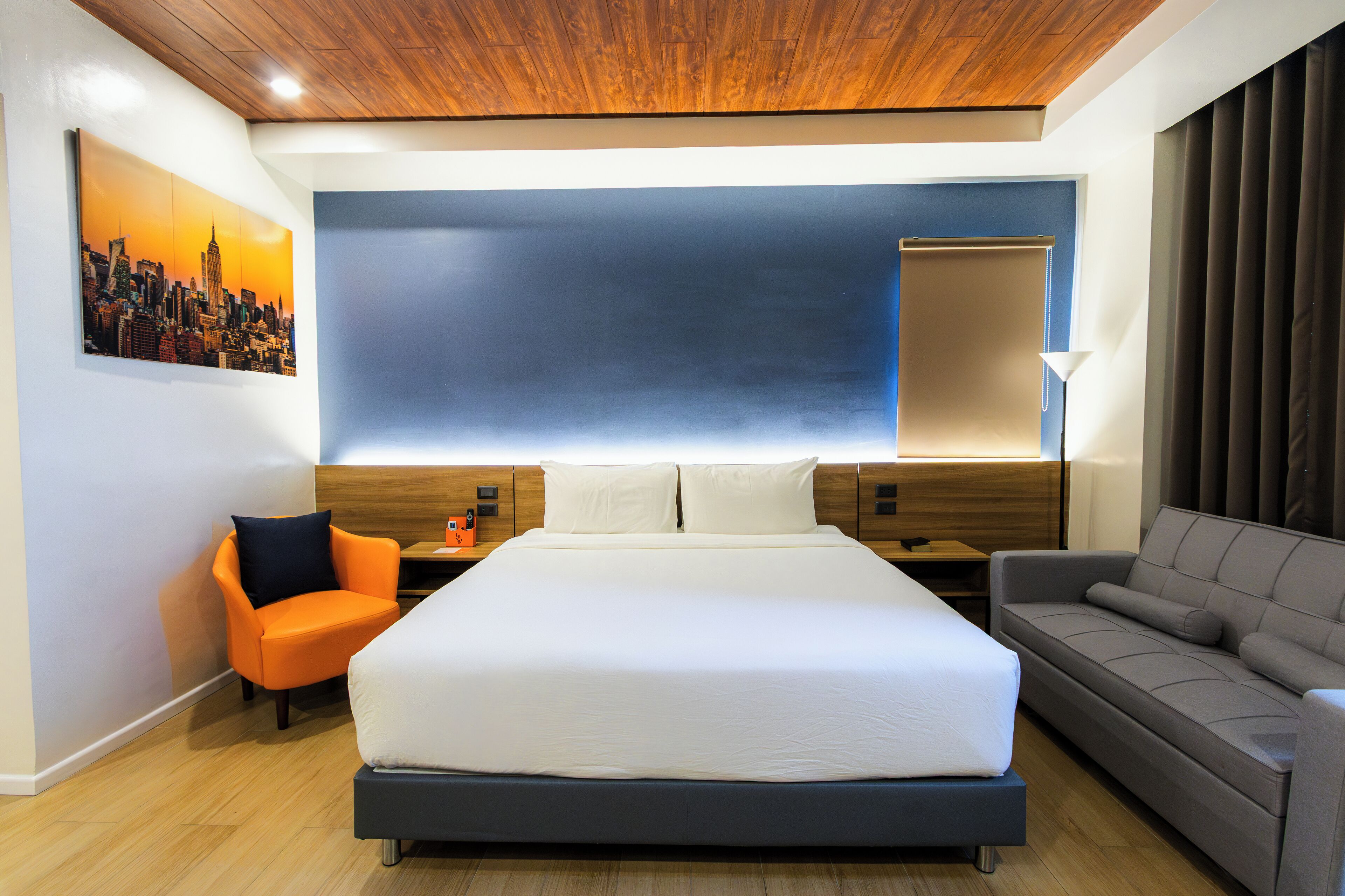 A modern hotel room with a large bed, a sofa, a chair, a nightstand, a lamp, and a cityscape painting on the wall.