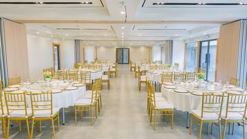 A spacious banquet hall with round tables and golden chairs, large windows on one side, and a modern ceiling design.