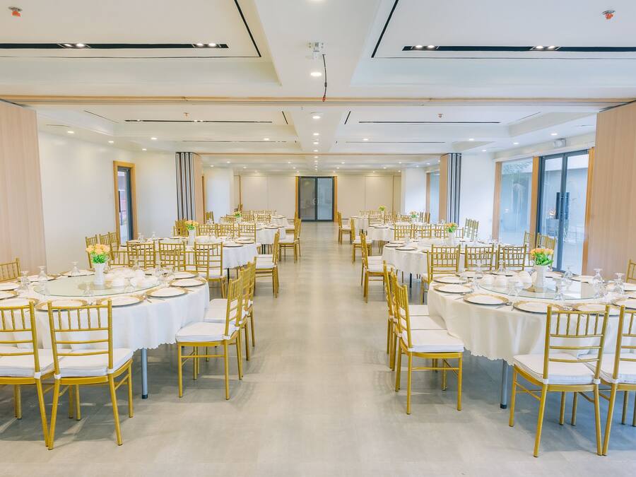A spacious banquet hall with round tables and golden chairs, large windows on one side, and a modern ceiling design.