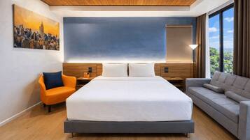 A modern hotel room with a large bed, a sofa, a chair, a cityscape painting, and a view of greenery.
