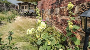 A brick building with a table and chairs outside, surrounded by greenery and flowers. - Red Grouse Cottage (Guisborough)