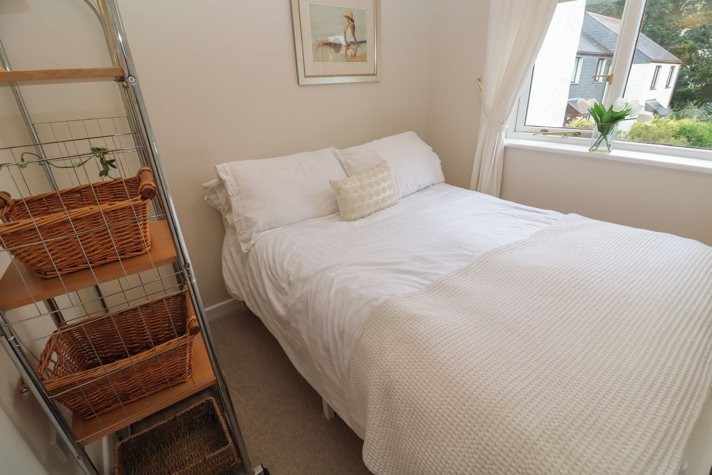 A neatly arranged bedroom with a bed, a window with a view of houses, and a metal rack with baskets.
