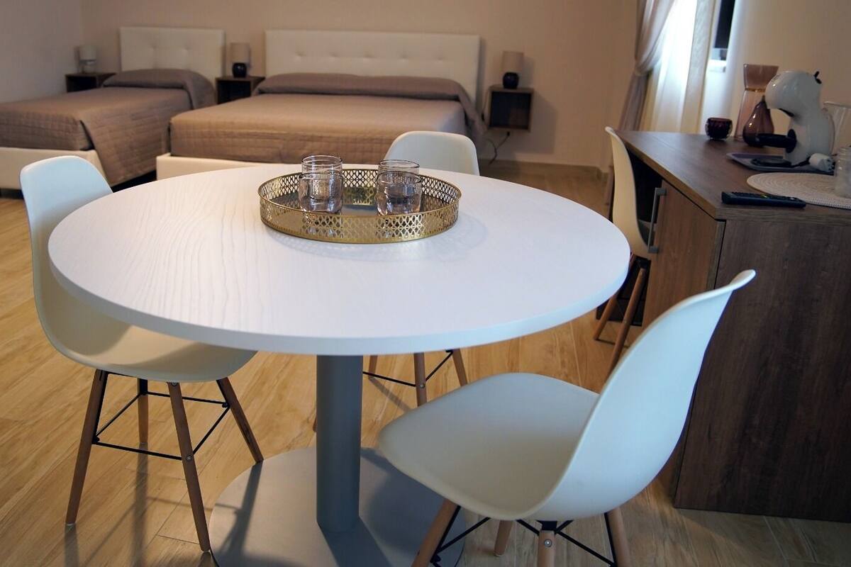 A modern hotel room with a round dining table, white chairs, a bed with a beige duvet, and a small side table with a lamp.