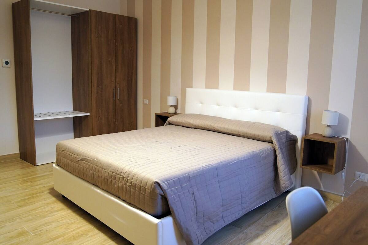 A bedroom with a bed, a wardrobe, a bedside table with a lamp, and a small wooden shelf.