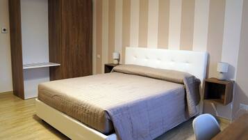 A bedroom with a bed, a wardrobe, a bedside table with a lamp, and a small wooden shelf.