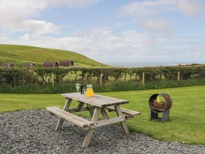 A picnic table with a bottle of wine, a glass, and a pitcher of juice, set on a gravel area with a view of a grassy hill and a body of water in the background. - Forest (Dalton-in-Furness)