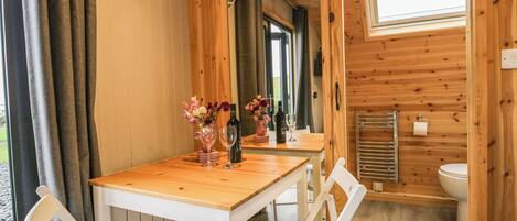 A wooden cabin with a dining area featuring a table, chairs, and a mirror. There is a window and a door leading to a bathroom.