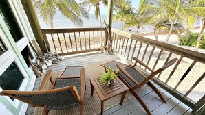 A wooden deck with a table and chairs, overlooking a beach with palm trees.